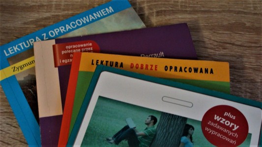 Książki ułożone na biurku w formę wachlarza (jedna na drugiej). Widoczne fragmenty okładek m.in. z napisem "lektura z opracowaniem".. Kliknij, aby powiększyć zdjęcie.